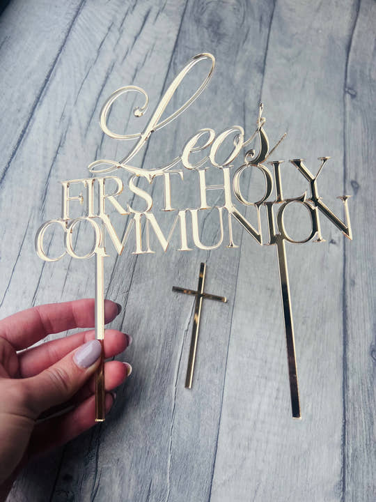 Gold 'Leo's First Holy Communion' cake topper with a cross on a wooden surface.