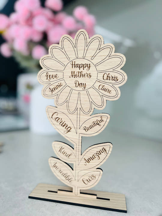 A freestanding wooden flower-shaped plaque with personalized engravings, intended as a Mother's Day card, displayed on a table.