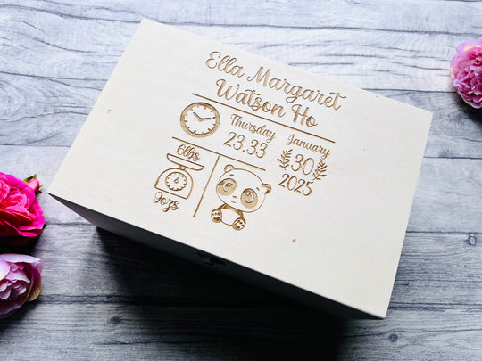 A white birch wood memory box with gold effect personalized engravings, placed on a grey surface with pink flowers around it.