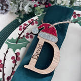 Load image into Gallery viewer, A personalized oak veneer name place table setting with 'D' for David, decorated with a red and white Santa hat, hanging from a green ribbon on a Christmas-themed plate.