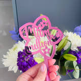 Load image into Gallery viewer, A hand holding a pink acrylic flower insert with the words 'Mum you billion' cut out of it, set against the backdrop of a bouquet of purple and white flowers.