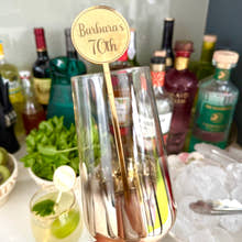 Load image into Gallery viewer, Rose gold cocktail glass with a 'Barbara's 70th' stirrer held by a hand, with bottles and drinks in the background.