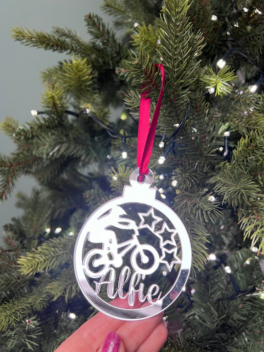 A hand holding a silver-colored Christmas tree decoration with a BMX bike and stars design, with a red hanging ribbon and lights in the background.