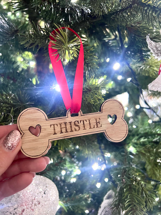 A personalised wooden bone-shaped decoration with the name 'Tristle' engraved on it, hanging from a red ribbon. It is displayed against a blurred Christmas tree background.