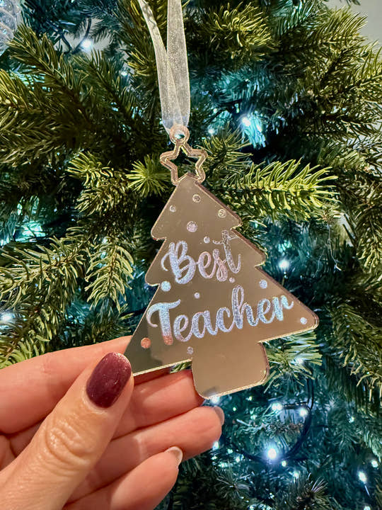Decorative ornament with 'Best Teacher' text held in front of a Christmas tree.