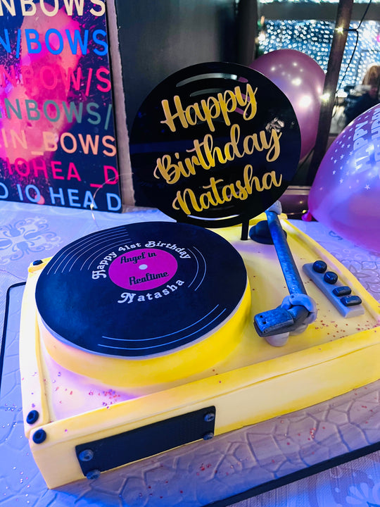 A cake topper featuring a black acrylic record design with customizable text in yellow and pink acrylic letters, placed on a cake with a birthday message.