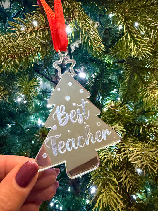 Decorative ornament shaped like a tree with 'Best Teacher' text, held in front of a Christmas tree.