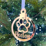 Load image into Gallery viewer, A gold-colored Christmas tree decoration in the shape of a paw print with the name 'Hector' engraved on it, hanging from a tree with lights and needles in the background.