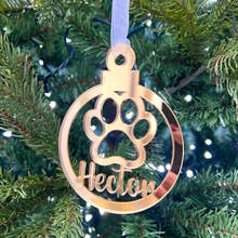 Load image into Gallery viewer, A gold-colored Christmas tree decoration in the shape of a paw print with the name 'Hector' engraved on it, hanging from a tree with lights and needles in the background.