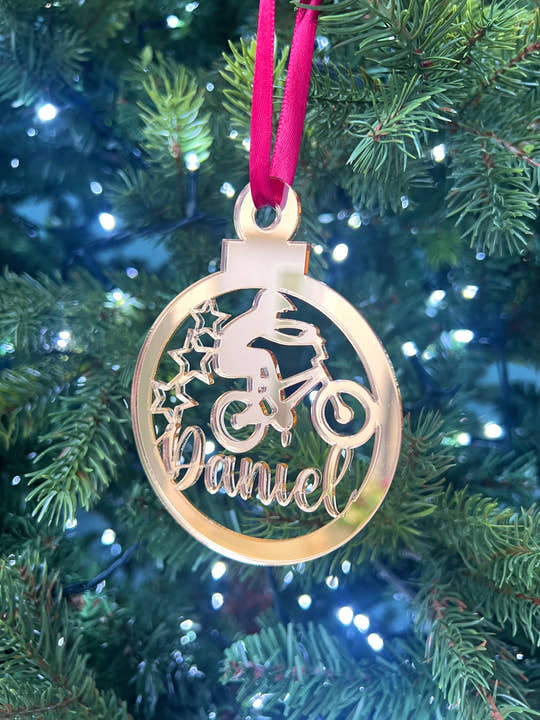 A gold-colored Christmas tree decoration featuring the text 'DANIEL' and a representation of a BMX bike, hanging from a red ribbon on a Christmas tree with green needles and decoration lights.
