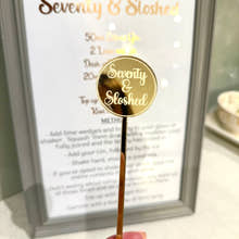 Load image into Gallery viewer, Gold cocktail stirrer with 'Seventy & Sloshed' branding held in front of a framed recipe card.