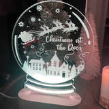 Load image into Gallery viewer, A clear acrylic Christmas snowglobe with a laser cut and engraved design featuring a reindeer, house, and Christmas tree, with a personalized text 'Christmas at the Door'.