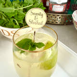 Load image into Gallery viewer, Glass of cocktail with mint leaves and a 'Seventy & Sloshed' stirrer, surrounded by bottles and ingredients on a countertop.