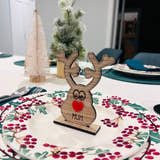Load image into Gallery viewer, Wooden reindeer place name holder with red nose and engraved name “Mum” on a festive Christmas dinner table with holly plates and holiday decorations.