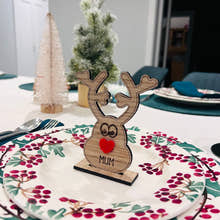 Load image into Gallery viewer, Wooden reindeer place name holder with red nose and engraved name “Mum” on a festive Christmas dinner table with holly plates and holiday decorations.