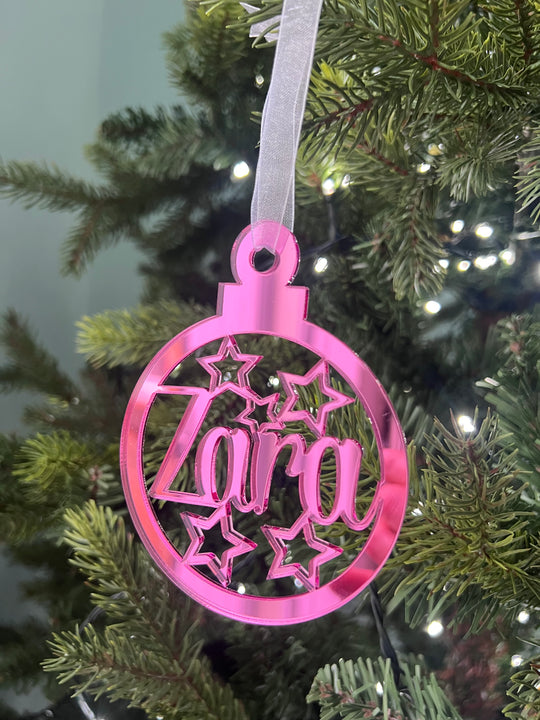 A pink, star-shaped Christmas tree decoration with the word 'Zara' engraved in the center, hanging from a tree with lights and greenery in the background.
