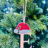 Load image into Gallery viewer, A decorative Christmas tree ornament in the shape of a Santa hat with the letter 'E' on it, hanging on a tree with lights and pine needles in the background.