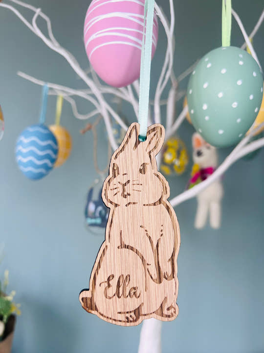 A wooden hanging bunny decoration with the name 'Ella' inscribed, displayed among colorful Easter eggs on a tree.