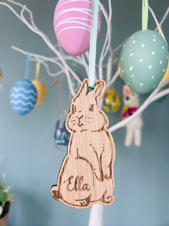 A wooden hanging bunny decoration with the name 'Ella' inscribed, displayed among colorful Easter eggs on a tree.