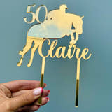 Load image into Gallery viewer, Close-up of personalised gold mirror acrylic horse cake topper featuring a horse rider silhouette, name and age.