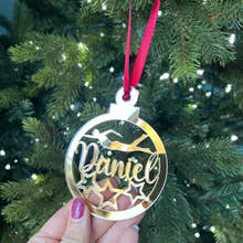 Load image into Gallery viewer, A hand holding a gold-colored Christmas tree decoration with the name 'Daniel' engraved on it, against a blurred background of a Christmas tree with lights.