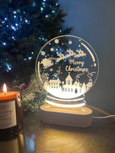 Load image into Gallery viewer, A clear acrylic snowglobe with a 'Merry Christmas' message, illuminated from within, placed on a table with a blurred Christmas tree and gifts in the background.