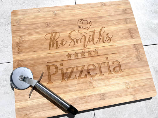 A wooden pizza board with laser engraved text that reads 'The Smiths Pizzeria' and includes a star symbol and a pizza cutter on the side.