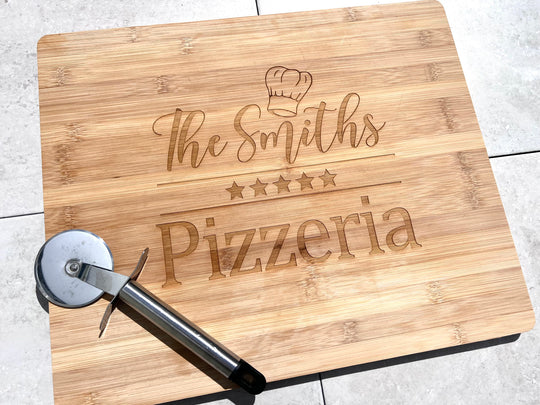 A wooden pizza board with laser engraved text that reads 'The Smiths Pizzeria' and includes a star symbol and a pizza cutter on the side.