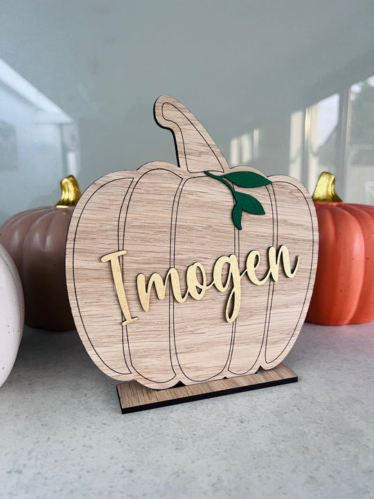 A wooden pumpkin-shaped decoration with 'Ingen' printed in metallic gold lettering, positioned on a stand.