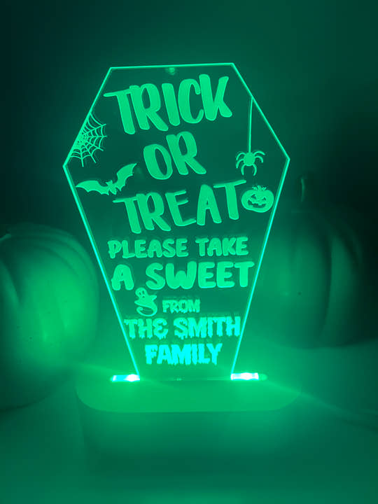 An LED light up stand with a personalized Halloween message 'Trick or Treat, Please Take a Sweet from The Smith Family' displayed on it. The stand is green and has a pumpkin-like shape with decorative elements and text.