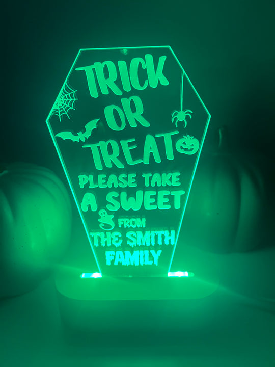 An LED light up stand with a personalized Halloween message 'Trick or Treat, Please Take a Sweet from The Smith Family' displayed on it. The stand is green and has a pumpkin-like shape with decorative elements and text.