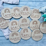 Load image into Gallery viewer, A set of wooden baby milestone discs with rainbow designs, each marked with different ages from one to twelve months, laid out next to a baby's clothing and a plush toy.