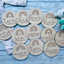 Load image into Gallery viewer, A set of wooden baby milestone discs with rainbow designs, each marked with different ages from one to twelve months, laid out next to a baby's clothing and a plush toy.