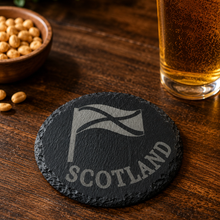 Load image into Gallery viewer, Slate coaster with 'Scotland' and tartan design on a wooden table with a glass of beer and bowl of nuts.