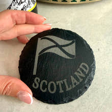 Load image into Gallery viewer, Round black slate coaster with 'Scotland' and a flag design, held by a hand on a light surface.