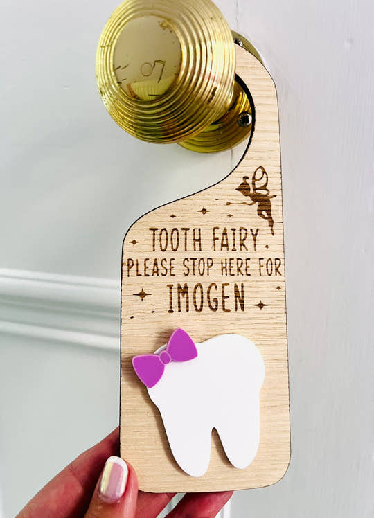 Personalised wooden Tooth Fairy door hanger with engraved text and white tooth shape with purple bow, hanging on a brass doorknob
