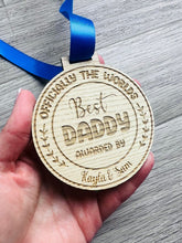Load image into Gallery viewer, A circular wooden medal engraved with the text “Officially the World’s Best Daddy,” “Awarded By Kayla & Sam,” featuring decorative leaves on the sides. The medal hangs from a royal blue satin ribbon