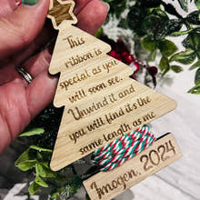 Load image into Gallery viewer, A personalized oak veneer Christmas tree decoration with engraved text, held in a hand, with a background of holly and decorative ribbon.
