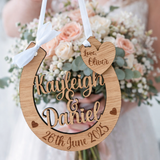 Load image into Gallery viewer, Bride holding personalised oak wooden wedding horseshoe engraved with names and date, hanging from white ribbon in front of bright colourful bridal bouquet, UK handmade wedding keepsake.
