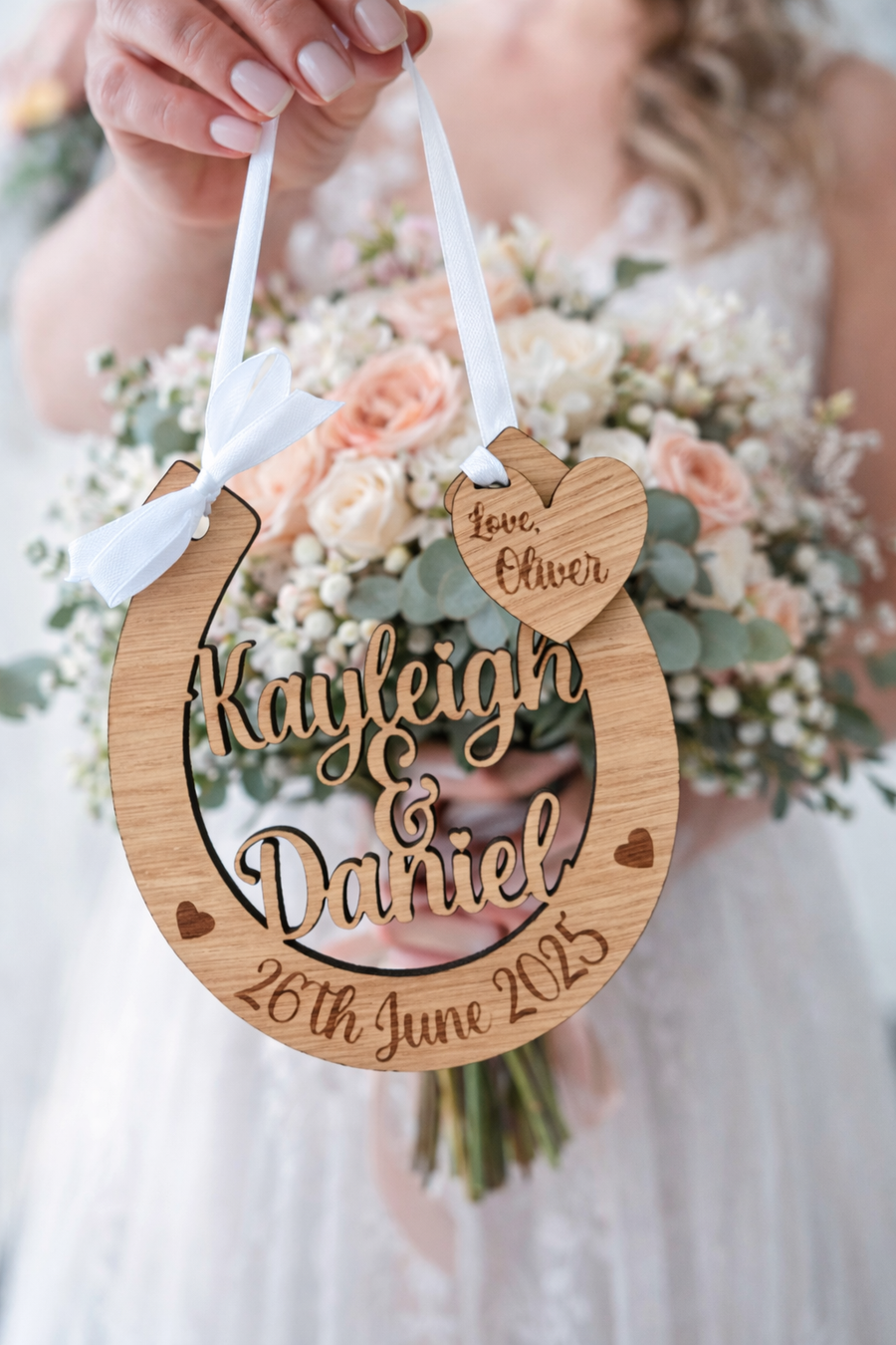 Bride holding personalised oak wooden wedding horseshoe engraved with names and date, hanging from white ribbon in front of bright colourful bridal bouquet, UK handmade wedding keepsake.