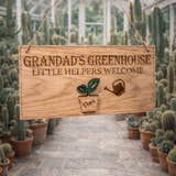 Load image into Gallery viewer, Personalised wooden sign reading “Grandad’s Greenhouse – Little Helpers Welcome” with a plant pot labelled Theo, hanging inside a bright cactus greenhouse with stone flooring.