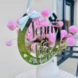 Load image into Gallery viewer, A mirrored horseshoe-shaped wedding decoration with the names "Jenn & Ian" and the date "12th April 2025" engraved. It has a light blue ribbon and bow for hanging, with pink flowers in the background.