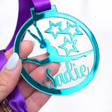 Load image into Gallery viewer, Personalised teal gymnastics medal with name Sadie, held in hand with purple ribbon, against blurred white hall background