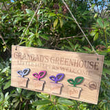 Load image into Gallery viewer, Personalised wooden sign reading “Grandad’s Greenhouse – Little Helpers Welcome” with a plant pot labelled with childrens name, hanging inside a bright garden