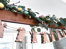 Load image into Gallery viewer, A wooden Easter Bunny Bunting hanging against a wall, featuring laser-cut bunny shapes with white pom-pom tails and decorative foliage.