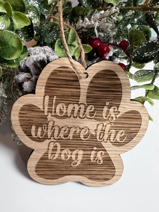 A wooden paw-shaped decoration with the text 'Home is where the Dog is' engraved on it, displayed against a backdrop of greenery and berries.