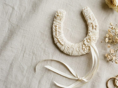 Lace-covered lucky wedding horseshoe with ribbon laid flat on linen beside dried flowers and wedding rings