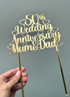 Gold acrylic cake topper with the words '50th Wedding Anniversary Mum & Dad' surrounded by hearts, designed for a golden anniversary celebration.