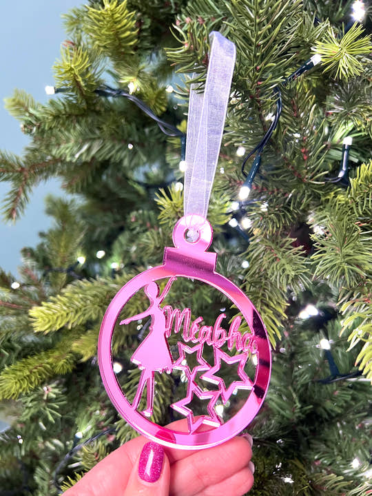 A custom-name Christmas tree decoration featuring a mirrored acrylic ballet dancer with a star, held against a blurred Christmas tree background. The decoration is in pink, with a name 'Mia' written on it.