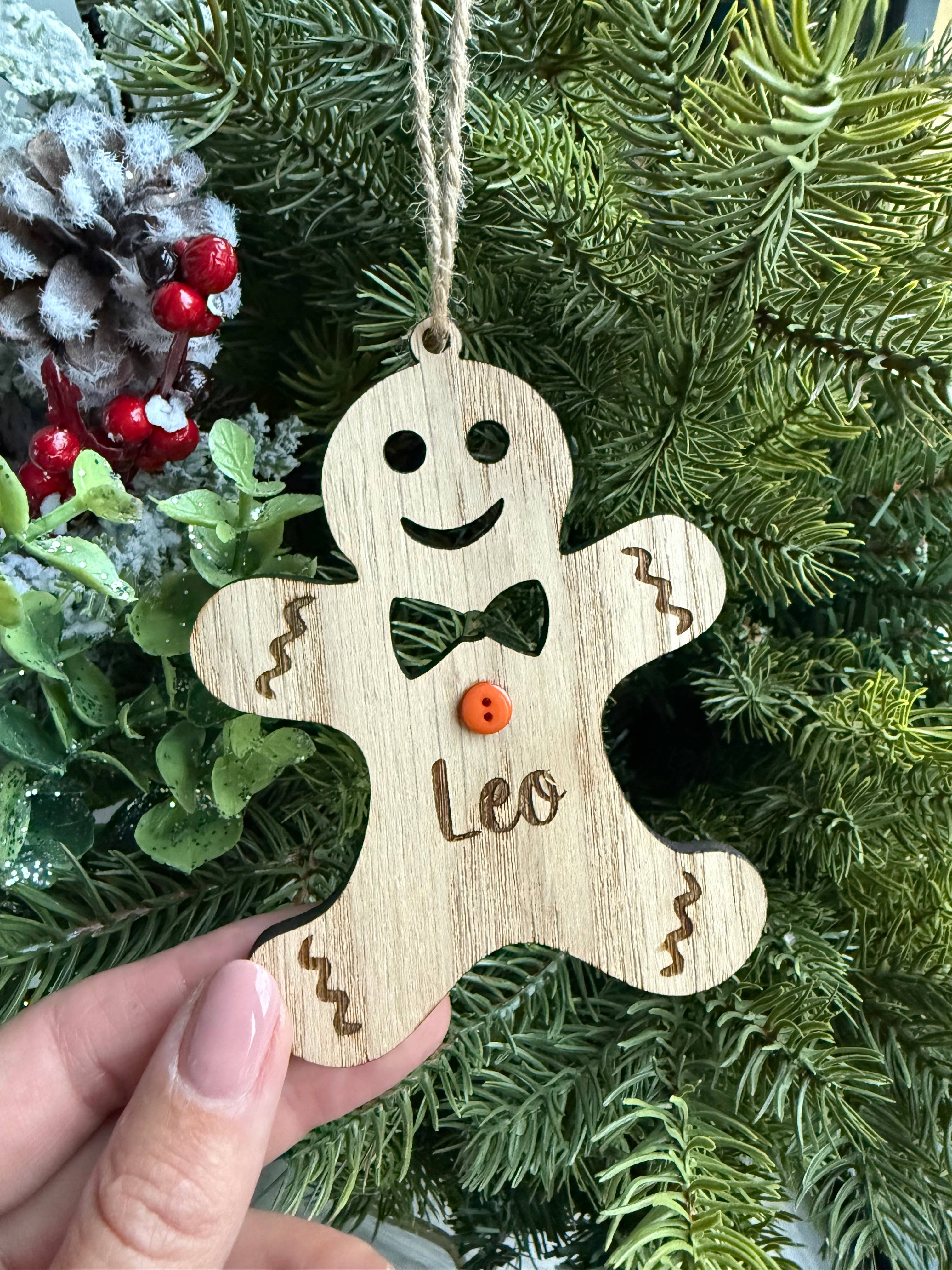 Wooden gingerbread man ornament with a face held in front of a Christmas tree.